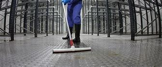 A person in blue overalls is cleaning the floor of a spacious warehouse with a broom