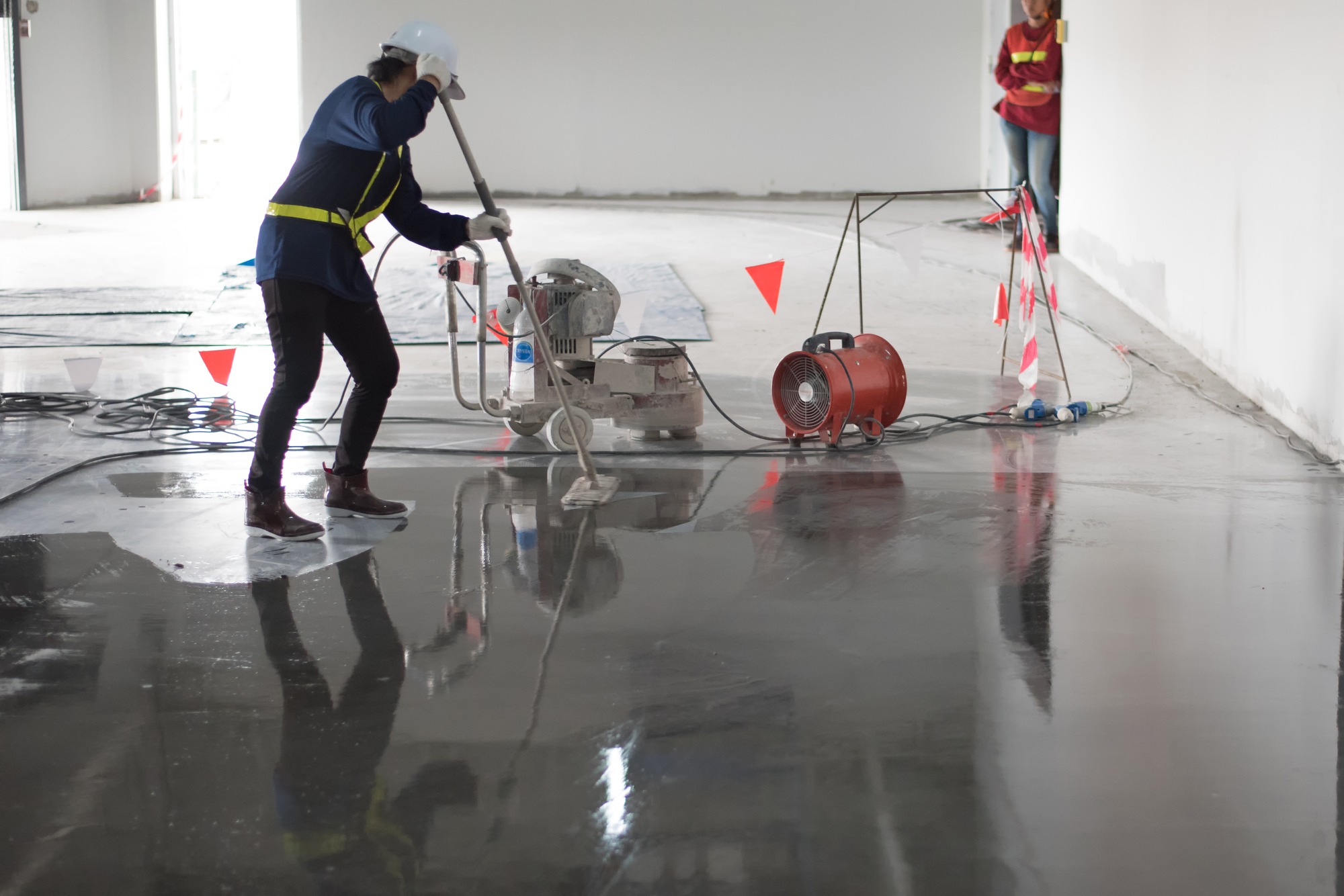 A worker in a hard hat and safety vest uses a mop on the floor at a construction site.