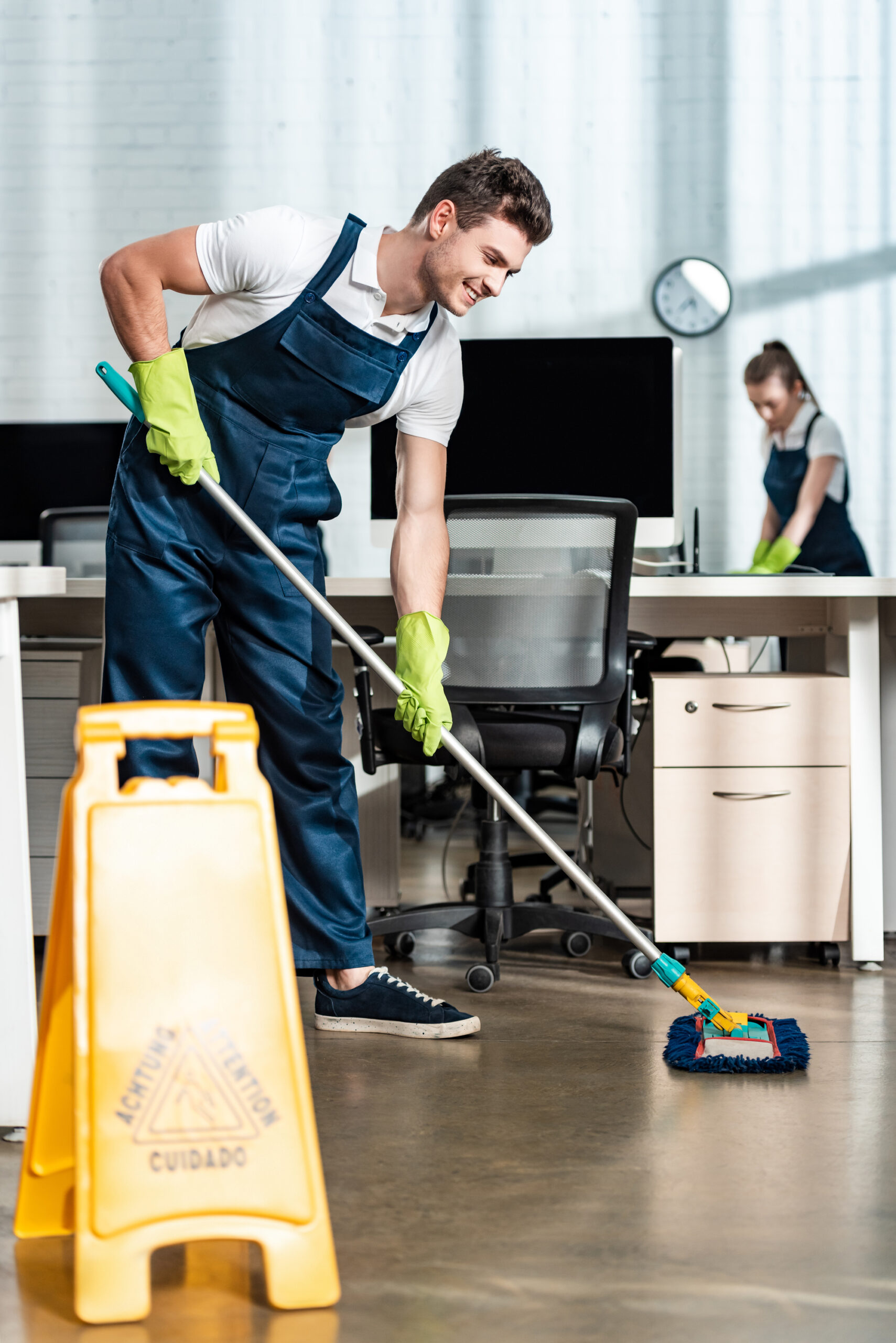 Cleaner washing floor with mop near colleague cleaning desk